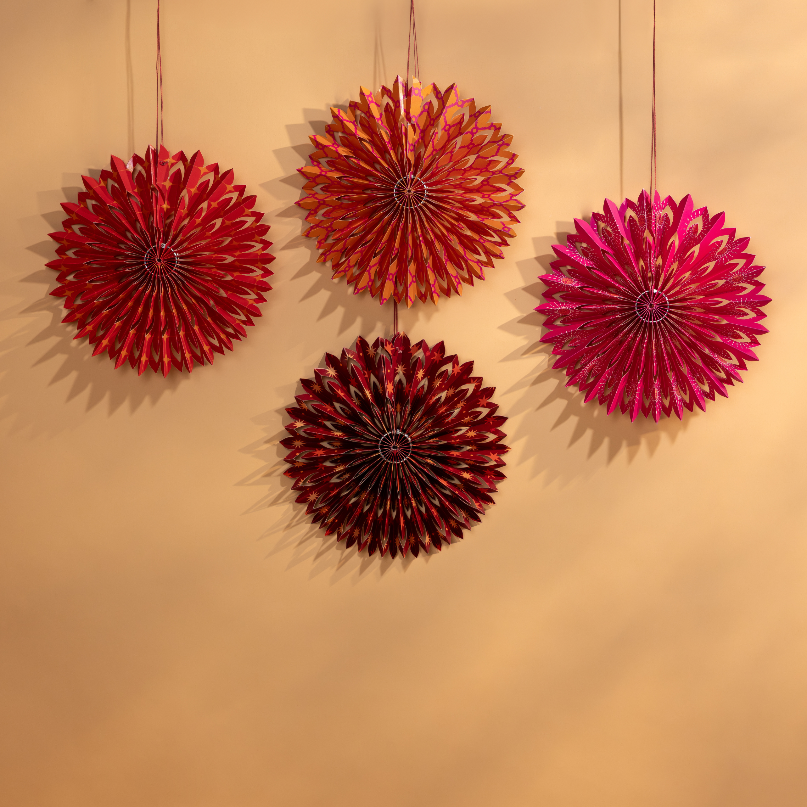 Four red and pink decorative fans hanging for Diwali decoration on a beige wall.