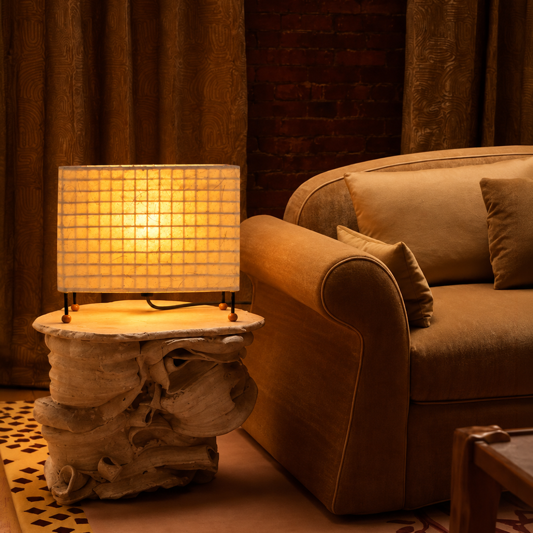 A warm glowing table lamp with a square grid shade sits on a sculptural wood side table next to a tan upholstered armchair in a cozy room setting.

