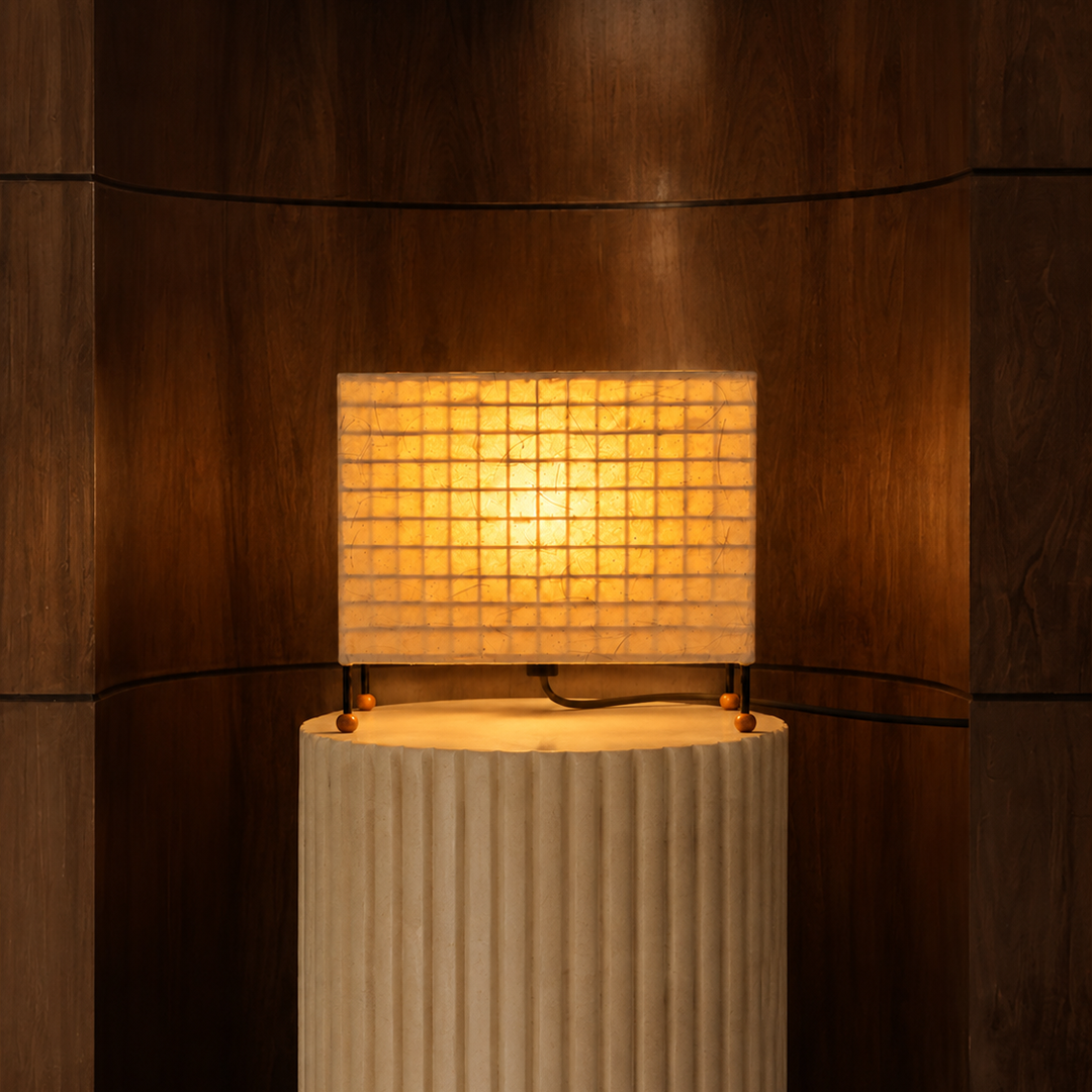 A modern table lamp with a glowing square grid shade resting on a ribbed cream pedestal against a dark wood-paneled background.

