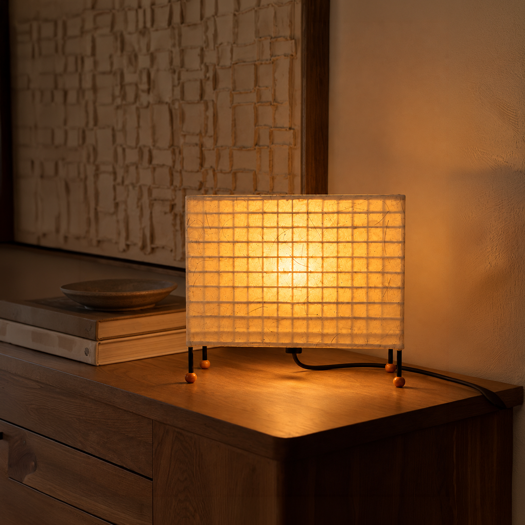 A rectangular table lamp with a glowing warm white grid-patterned shade and black legs, placed on a wooden dresser in a warmly lit room.