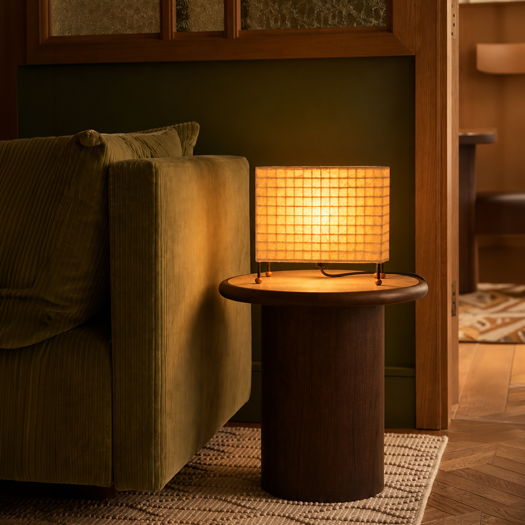 A warm glowing table lamp with a square grid shade sits on a round wooden side table next to an olive green armchair in a cozy room setting.

