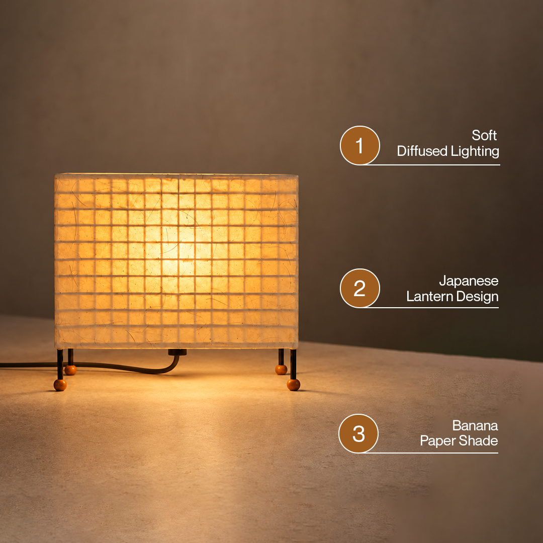 A Japanese-style table lamp with a square grid-paper shade glowing warm yellow, shown on a neutral surface against a dark gray background. Three circular callouts on the right describe features: soft diffused lighting, lantern design, and banana paper shade.