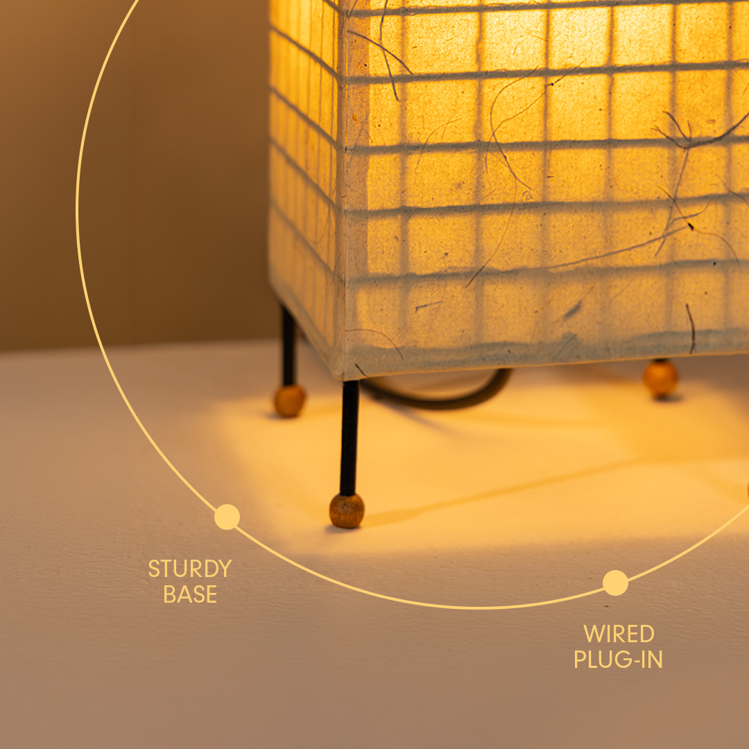 A close-up of a glowing table lamp with a square translucent shade, thin dark legs, and small wooden feet on a light surface. A faint circular graphic and text labels highlight the lamp's sturdy base and wired plug-in design.